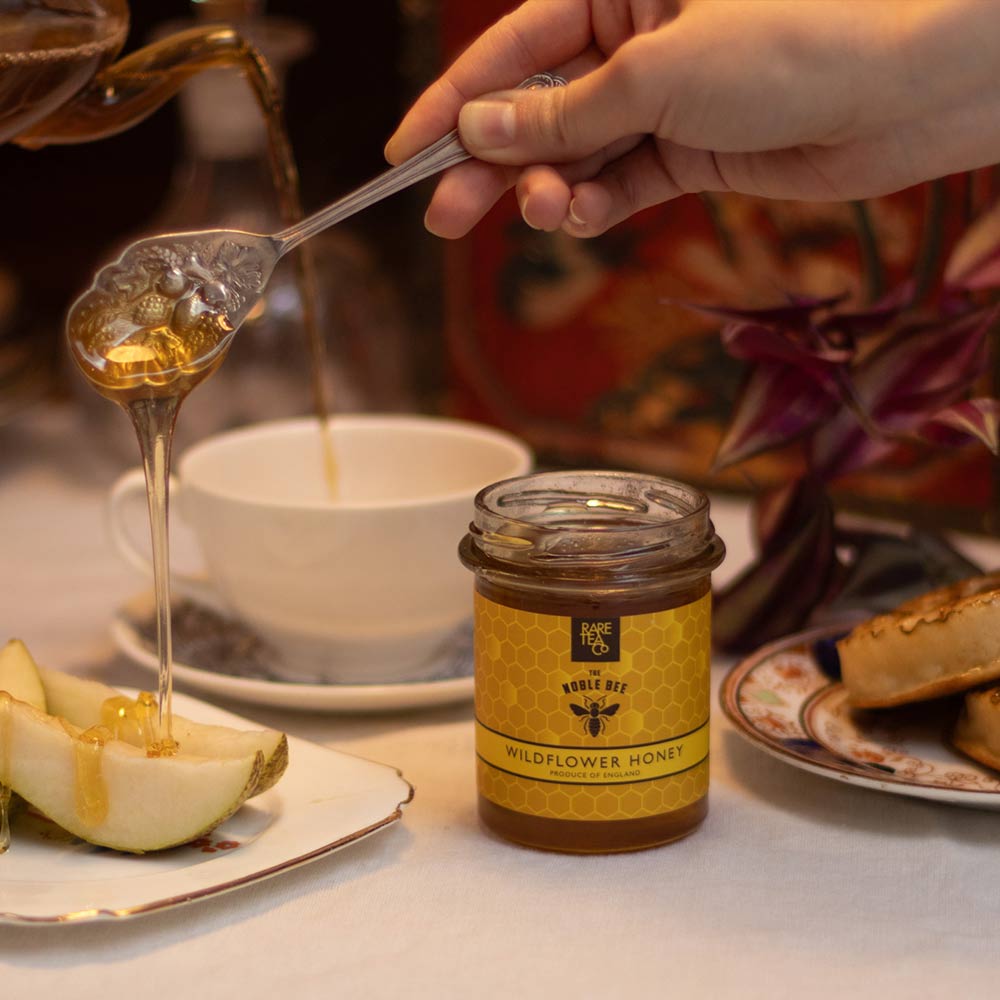 Person drizzling Rare Tea Wildflower Honey from a spoon onto fruit with the jar of honey and a tea cup in the background.