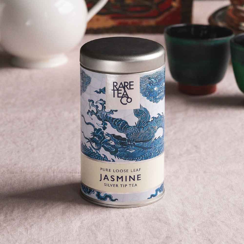 A tin of Rare Tea Company Silver Tip Jasmine tea on a white tablecloth with vintage teaware in the background.