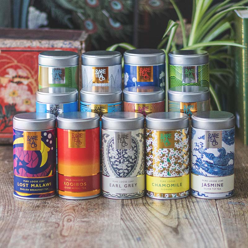 Collection of colourful Rare Tea Company tea tins on a wooden surface with plants in the background.