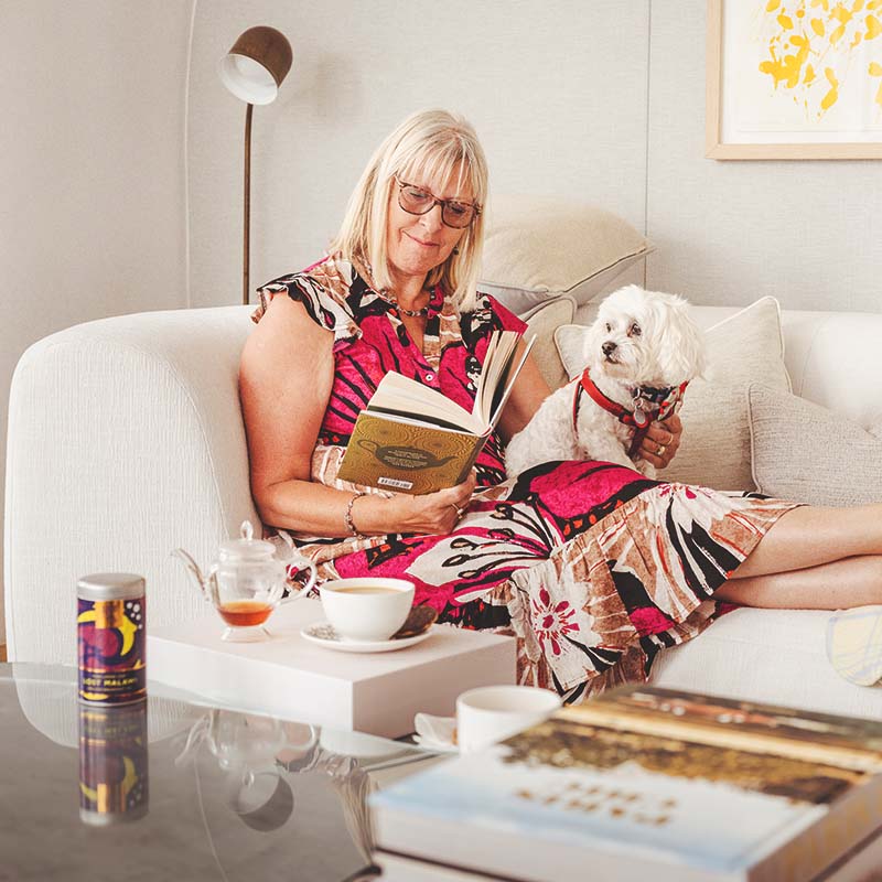 Woman reading a book with a small white dog on a couch in a cozy living room, with a teapot, tea cup and biscuit on a small table in front of her.