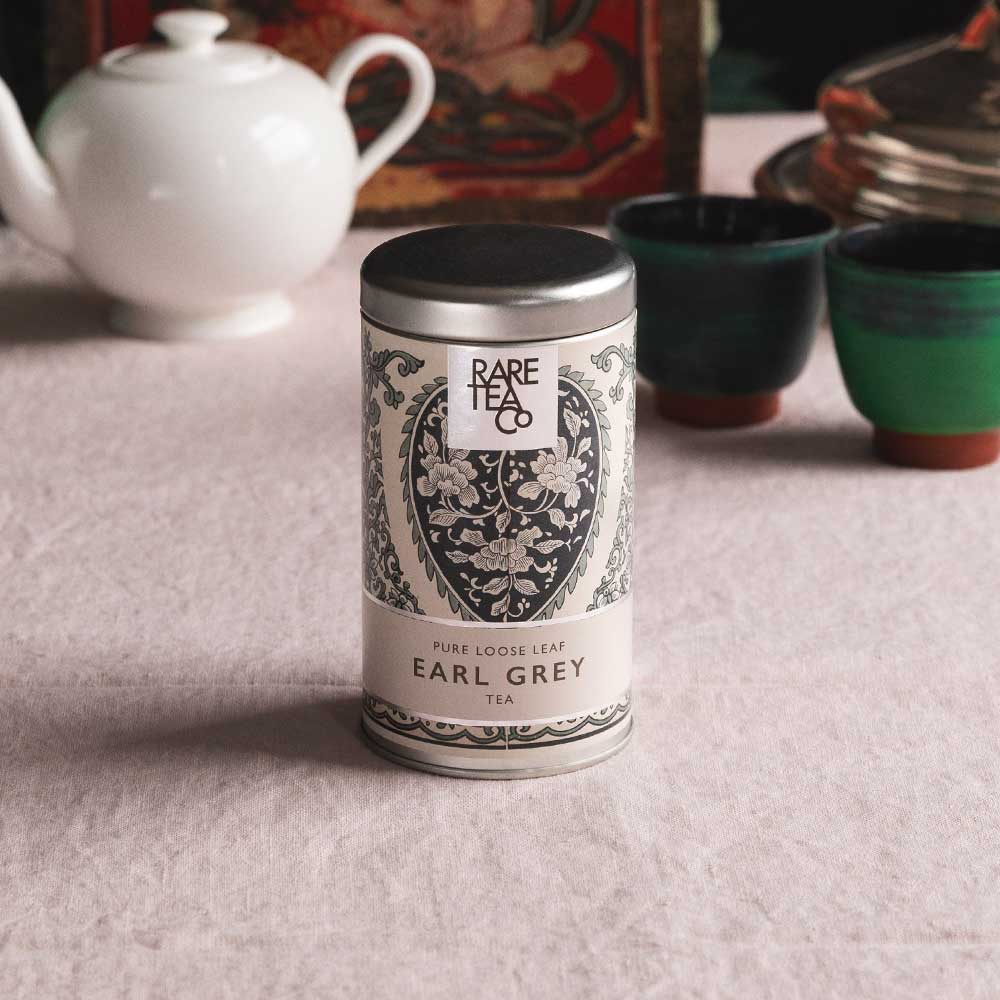 A tin of Rare Tea Company Rare Earl Grey on a white tablecloth with vintage teaware in the background.