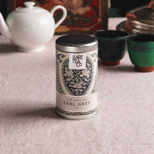 A tin of Rare Tea Company Rare Earl Grey on a white tablecloth with vintage teaware in the background.