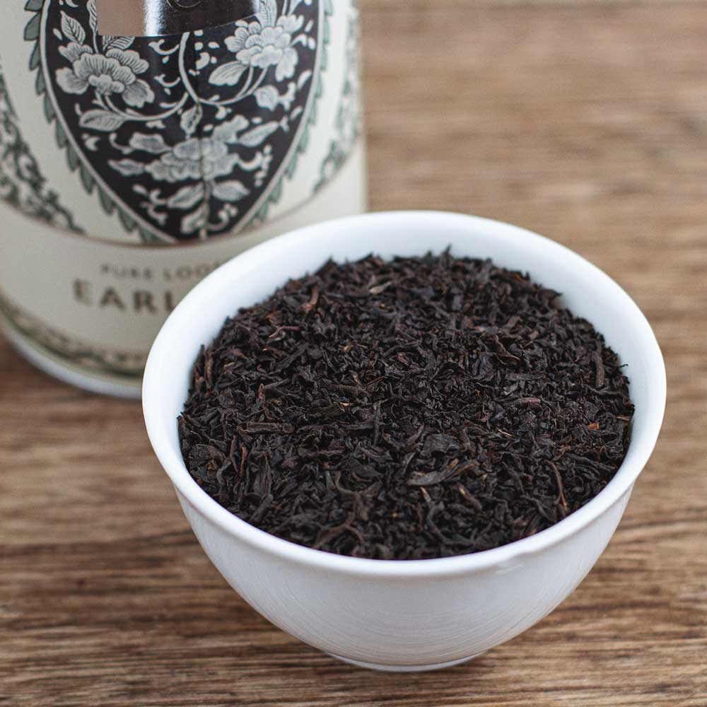 White bowl filled with Earl Grey tea leaves on a wooden surface, with a Rare Tea Company tea tin in the background.