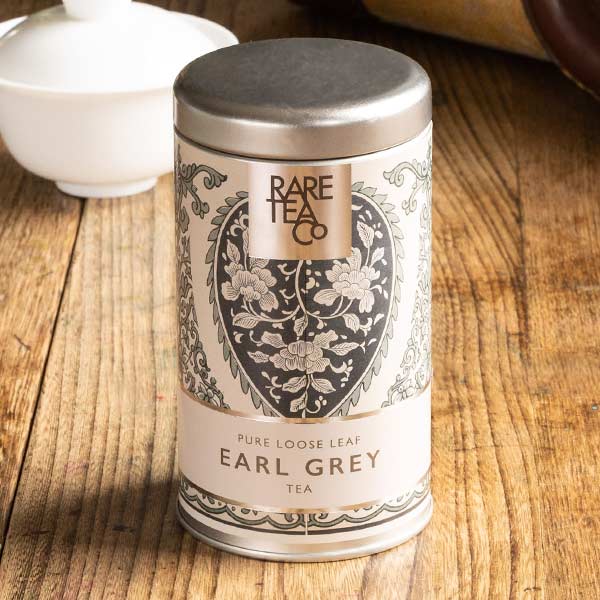 A tin of Rare Tea Company Rare Earl Grey tea on a wooden table with vintage teaware in the background.