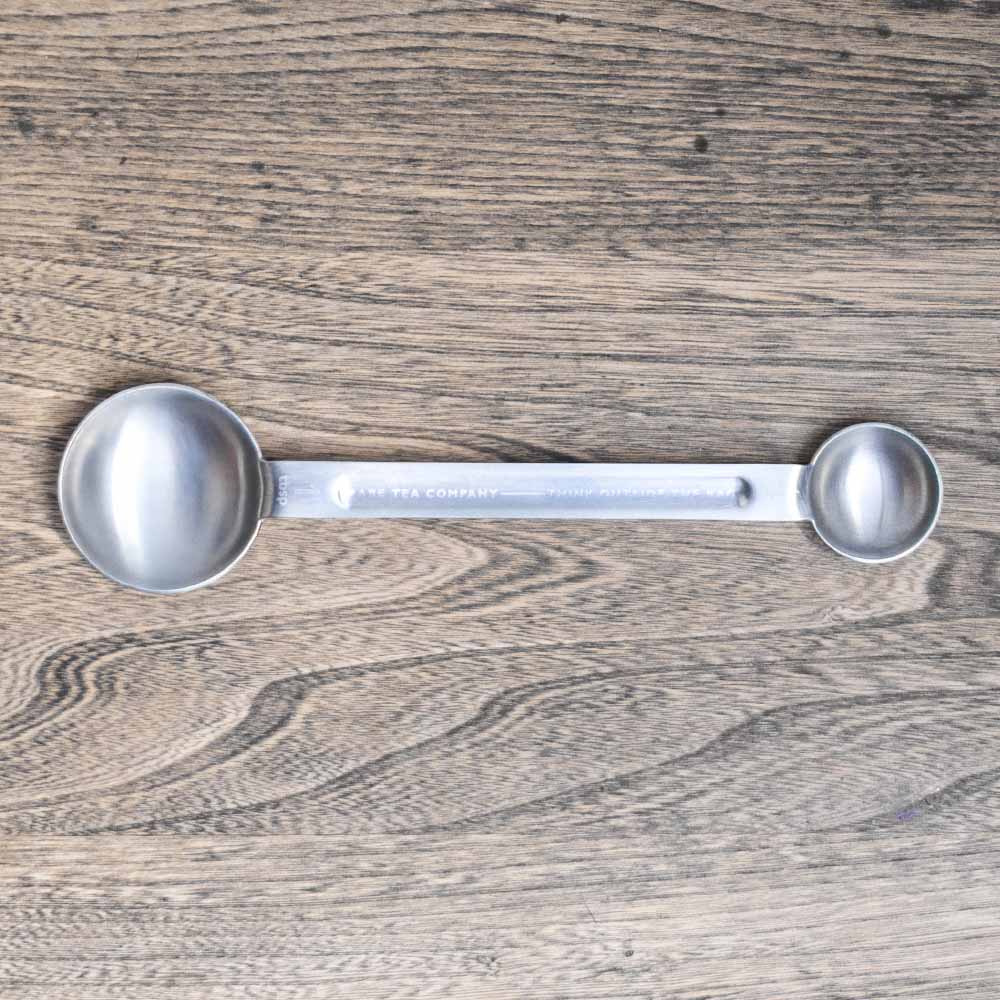Rare Tea double-ended metal measuring spoon on a wooden surface.