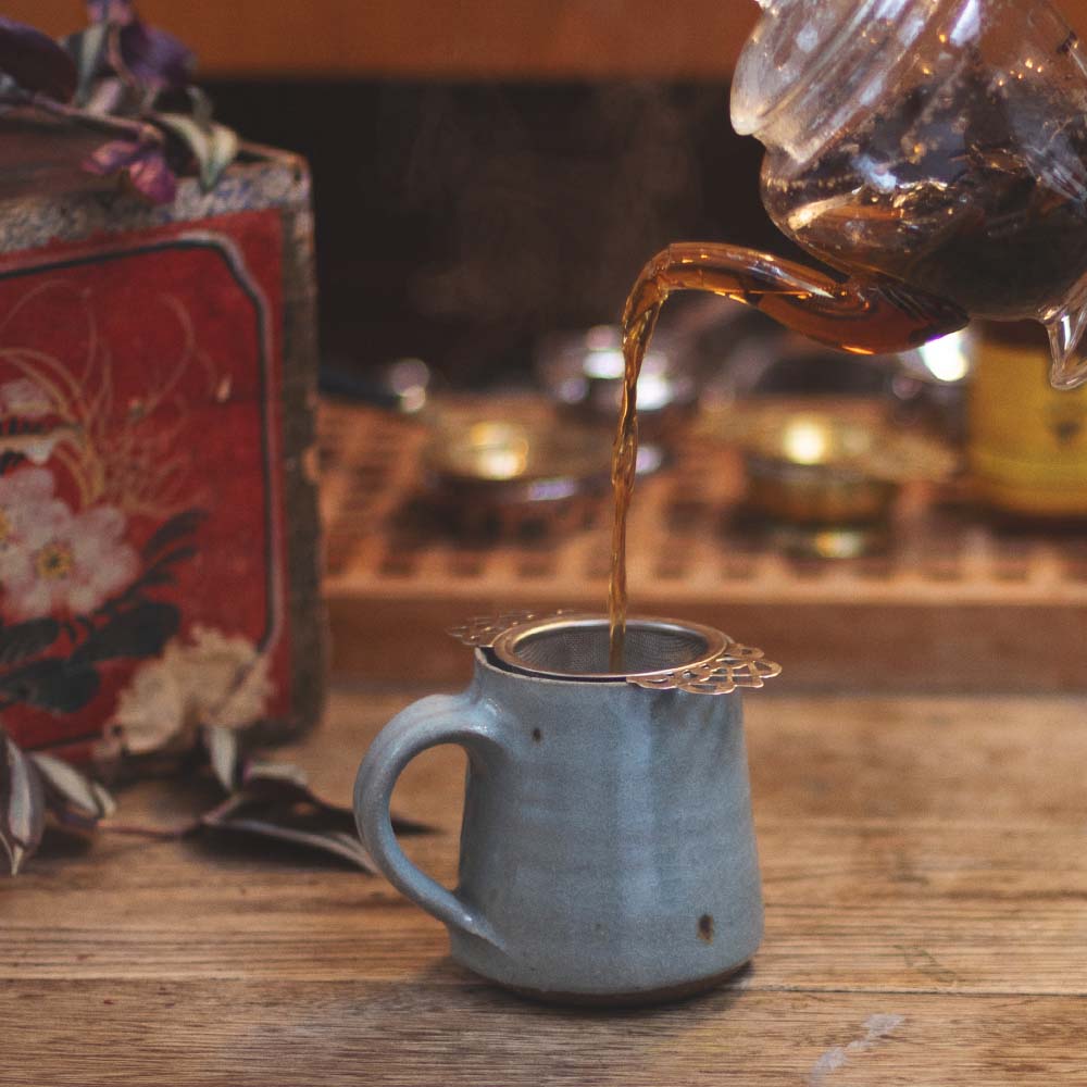 Tea being poured from a glass teapot into a ceramic mug on a wooden surface.