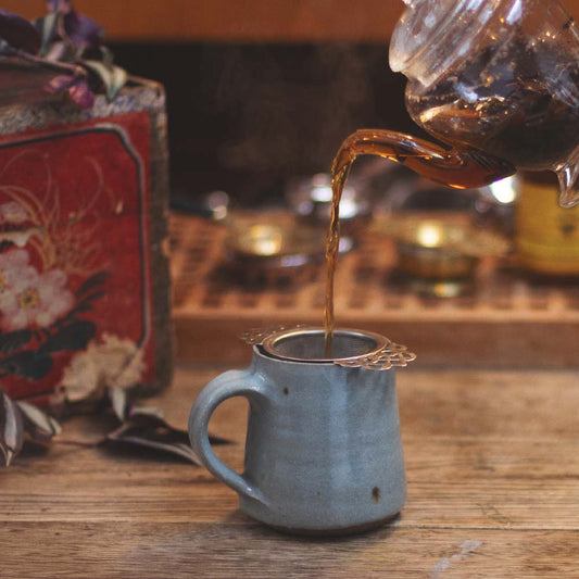 Tea being poured from a glass teapot into a ceramic mug on a wooden surface.