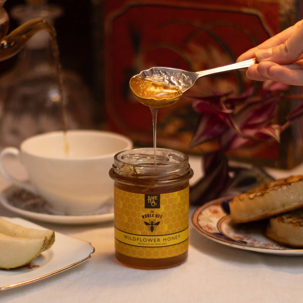 Rare Company Wildflower Honey being drizzled from a spoon into a jar labelled with a cup of tea and crumpets in the background.