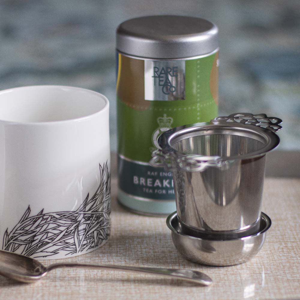A stainless steel loose tea infuser displayed alongside a mug, a teaspoon, and a tea canister, on a tray.