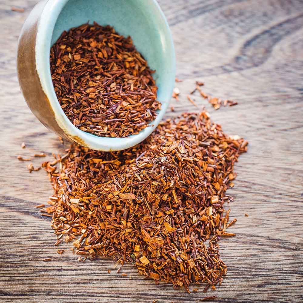 Dried rooibos tea leaves spilling out of a ceramic bowl on a wooden surface
